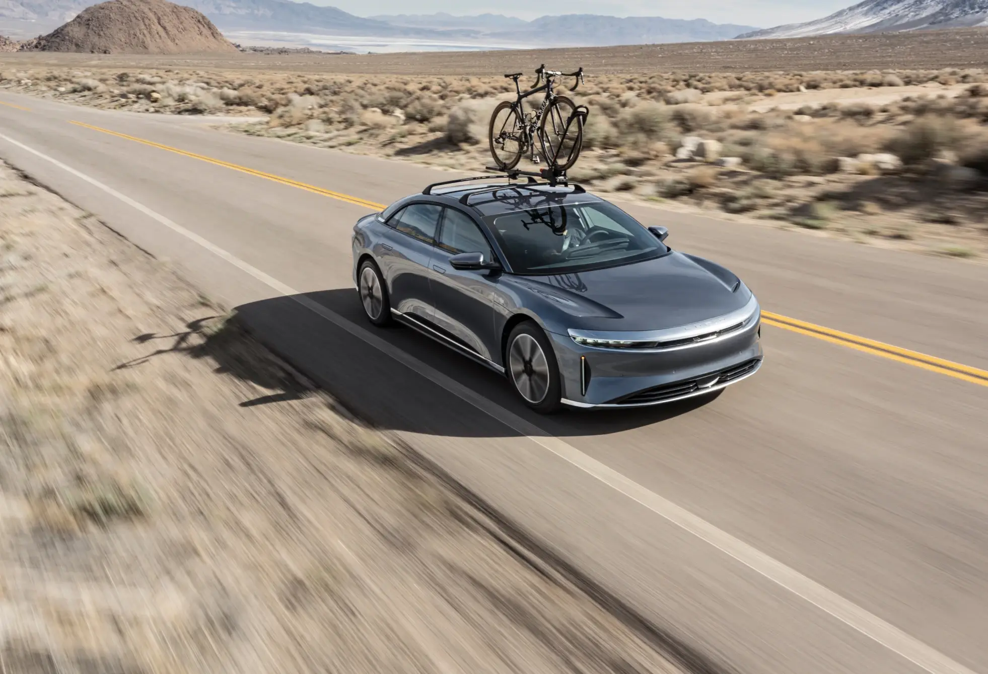 Een Lucid Air rijdt over een woestijnweg met twee fietsen op het dak, omringd door rotsachtig terrein en verre bergen.
