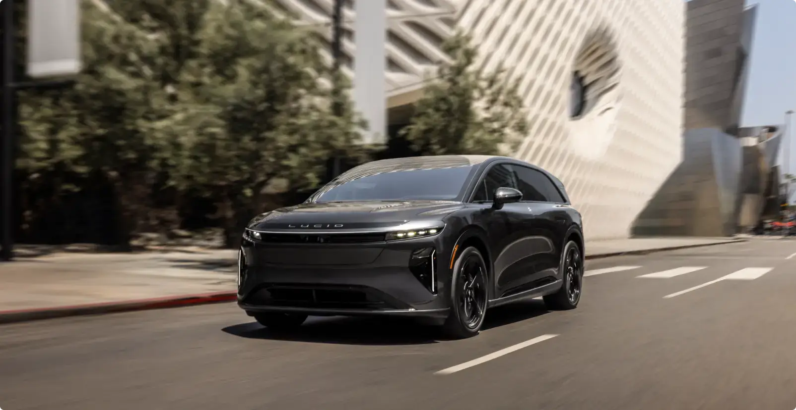 A sleek black Lucid electric SUV driving on a city street, with trees and a modern white building in the background.