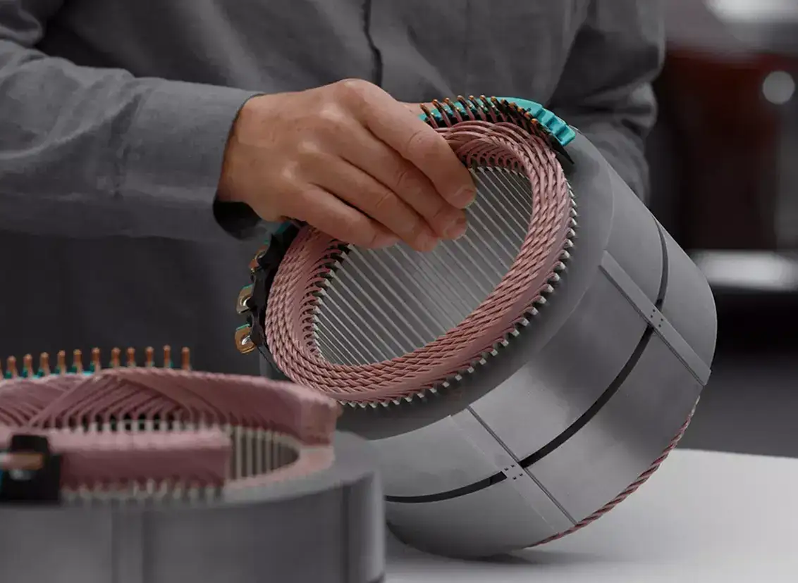 Hands assembling an electric motor with copper wire coils wrapped around a cylindrical metal stator in a manufacturing setting.