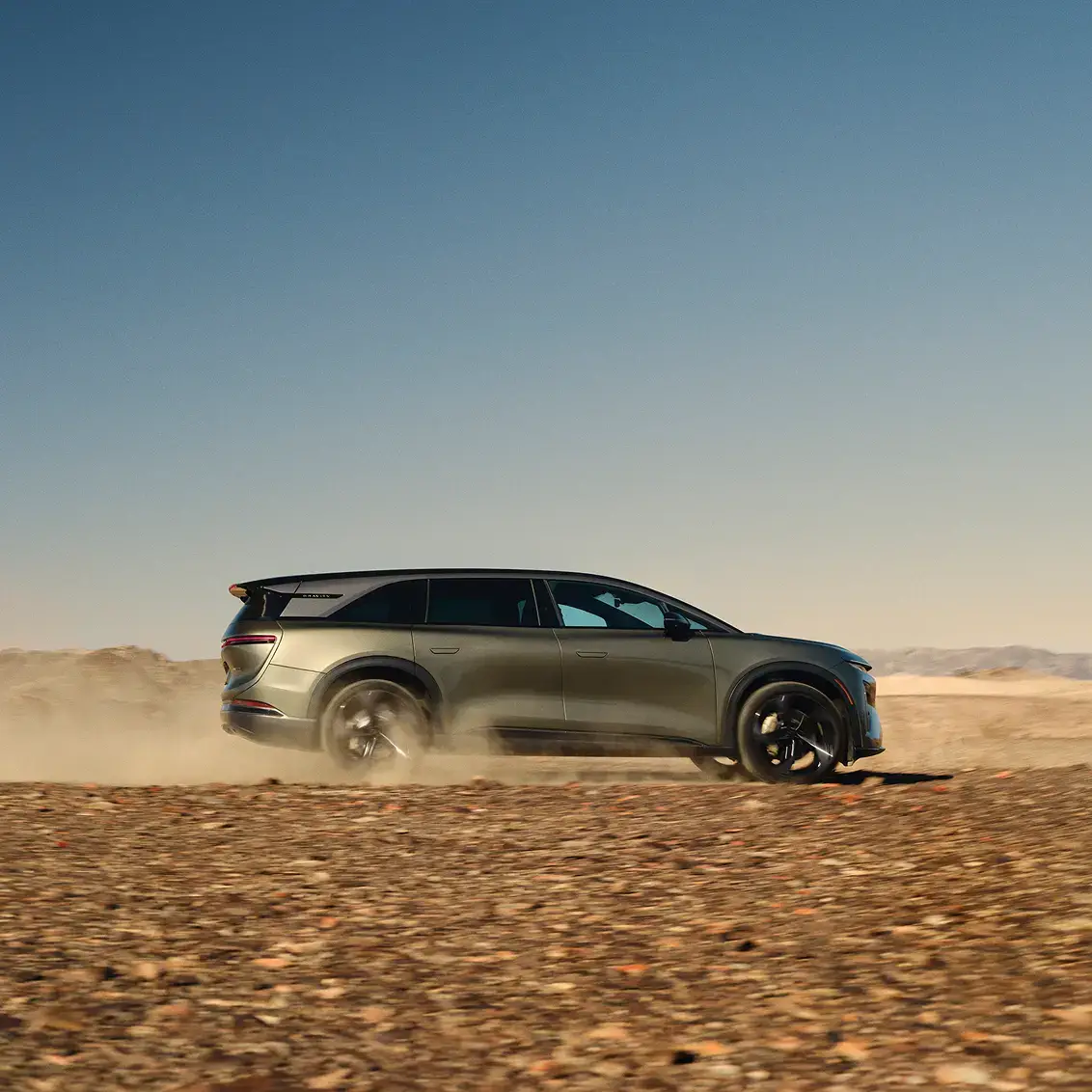 Lucid Gravity driving through a dusty desert landscape, kicking up dust, with a clear blue sky and sandy hills in the background.