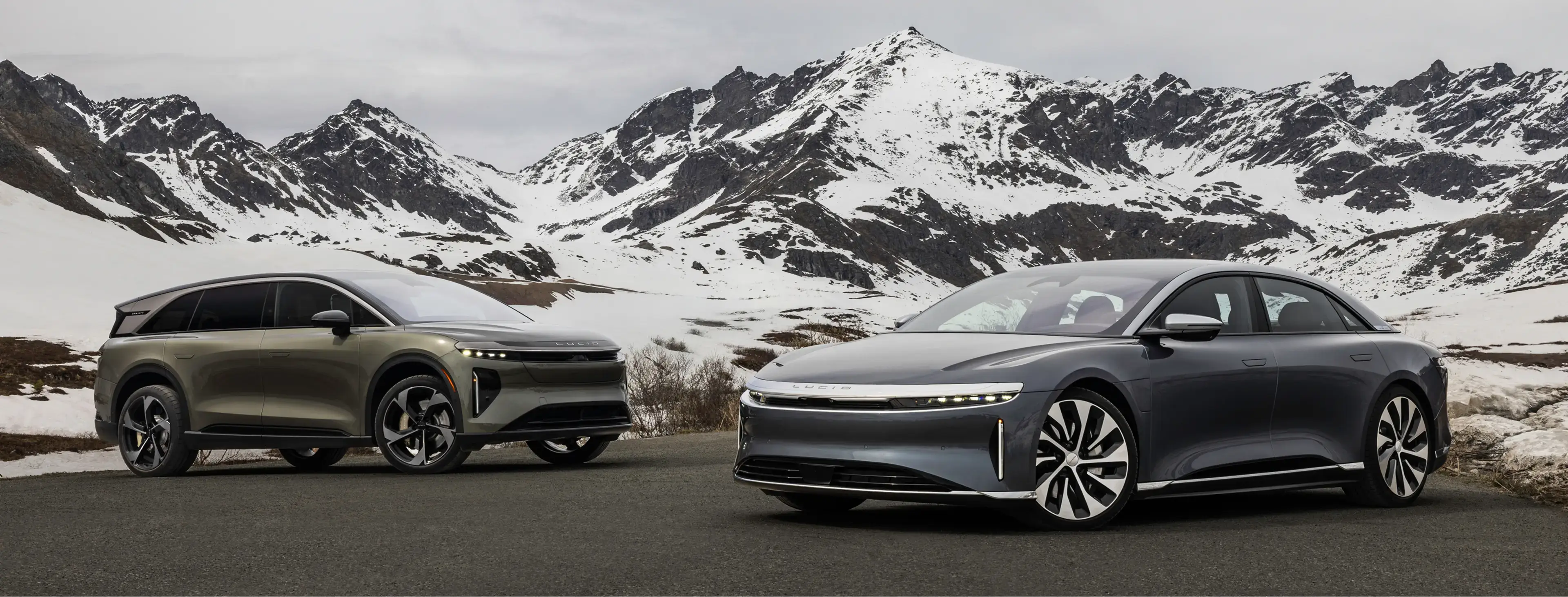 Two Lucid electric vehicles parked on a mountain road with snow-capped peaks in the background under an overcast sky.