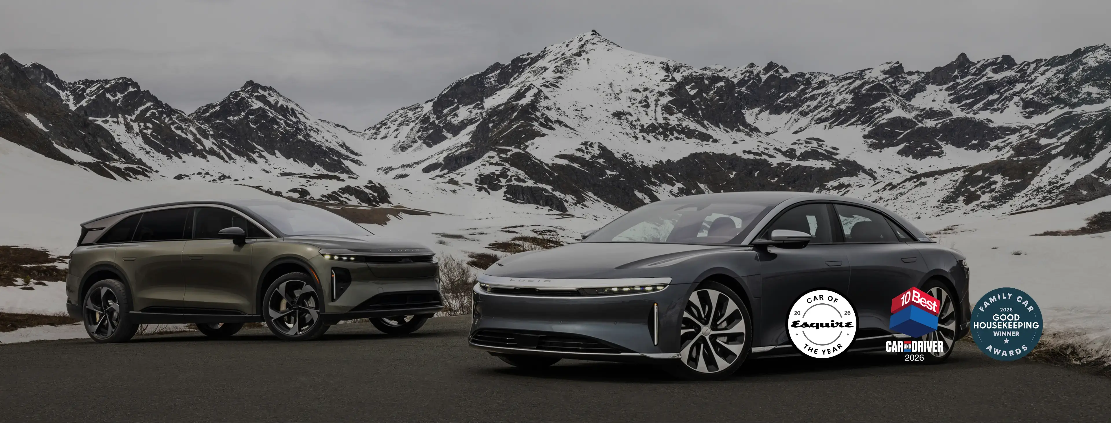 Two Lucid electric vehicles parked on a mountain road with snow-capped peaks in the background under an overcast sky.