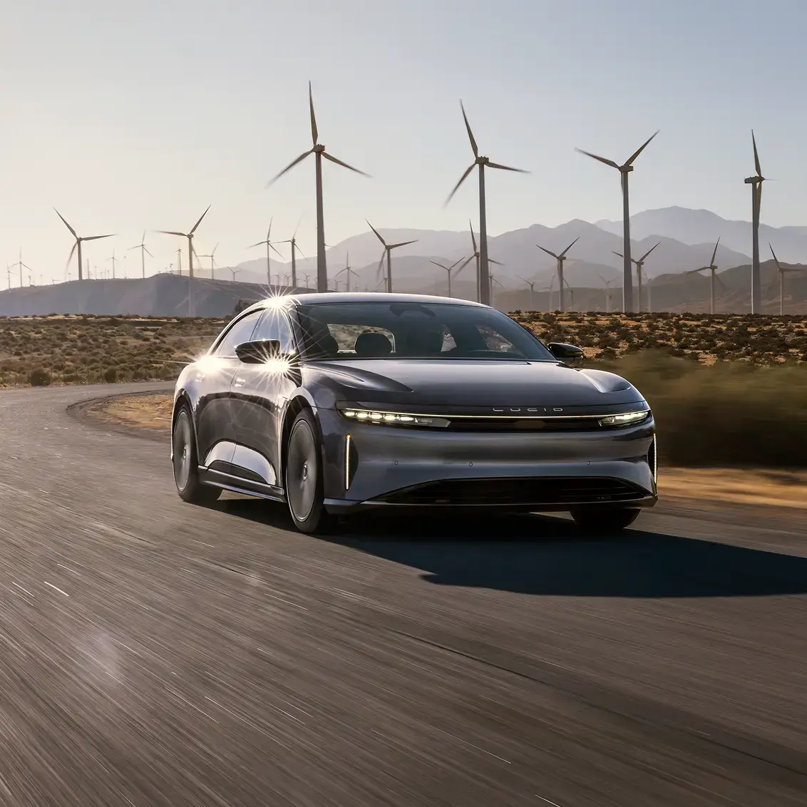 Sleek Lucid electric car driving on desert road with wind turbines and mountains in background at sunset.