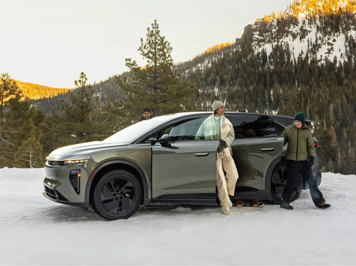 Des personnes avec un VUS Lucid Gravity dans les montagnes enneigées au coucher du soleil, avec des pins et des sommets recouverts de neige en arrière‑plan.