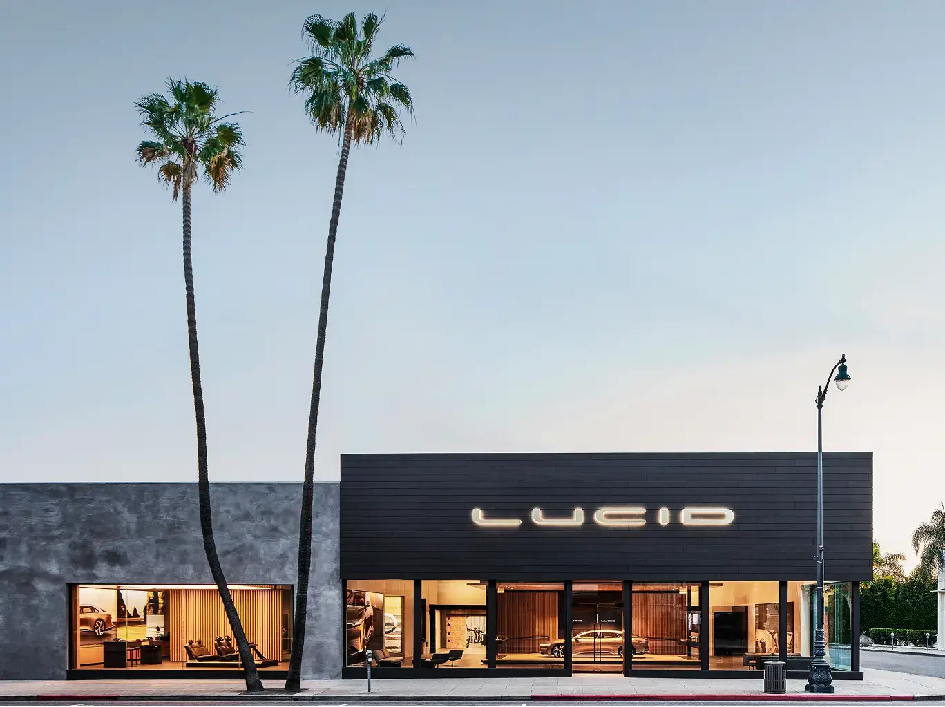 Lucid Motors showroom with illuminated sign on dark facade, tall palm trees against clear sky, and glass storefront.