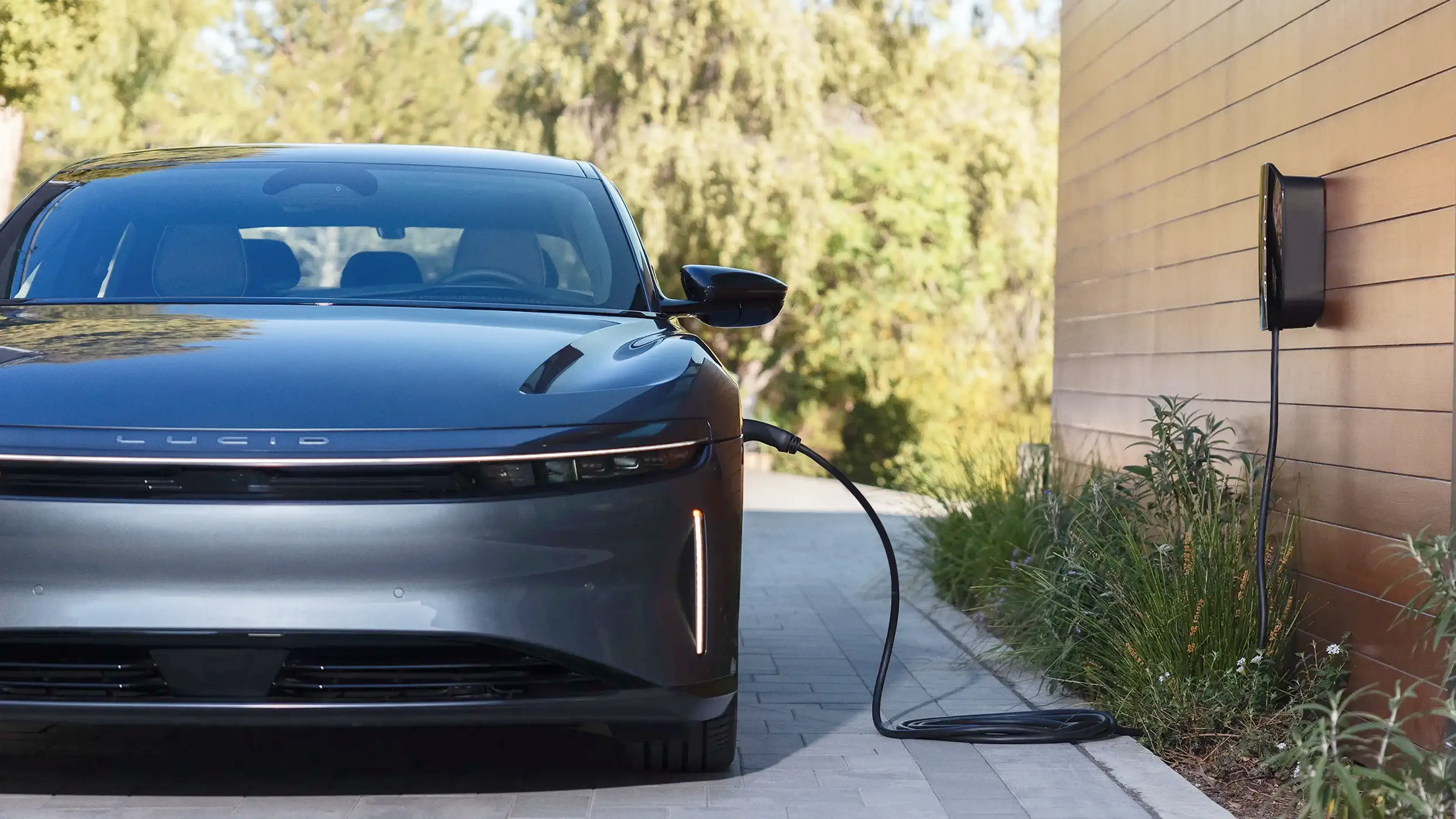 Electric car charging at home with a wall-mounted charger, sleek black vehicle parked on paved driveway beside greenery.