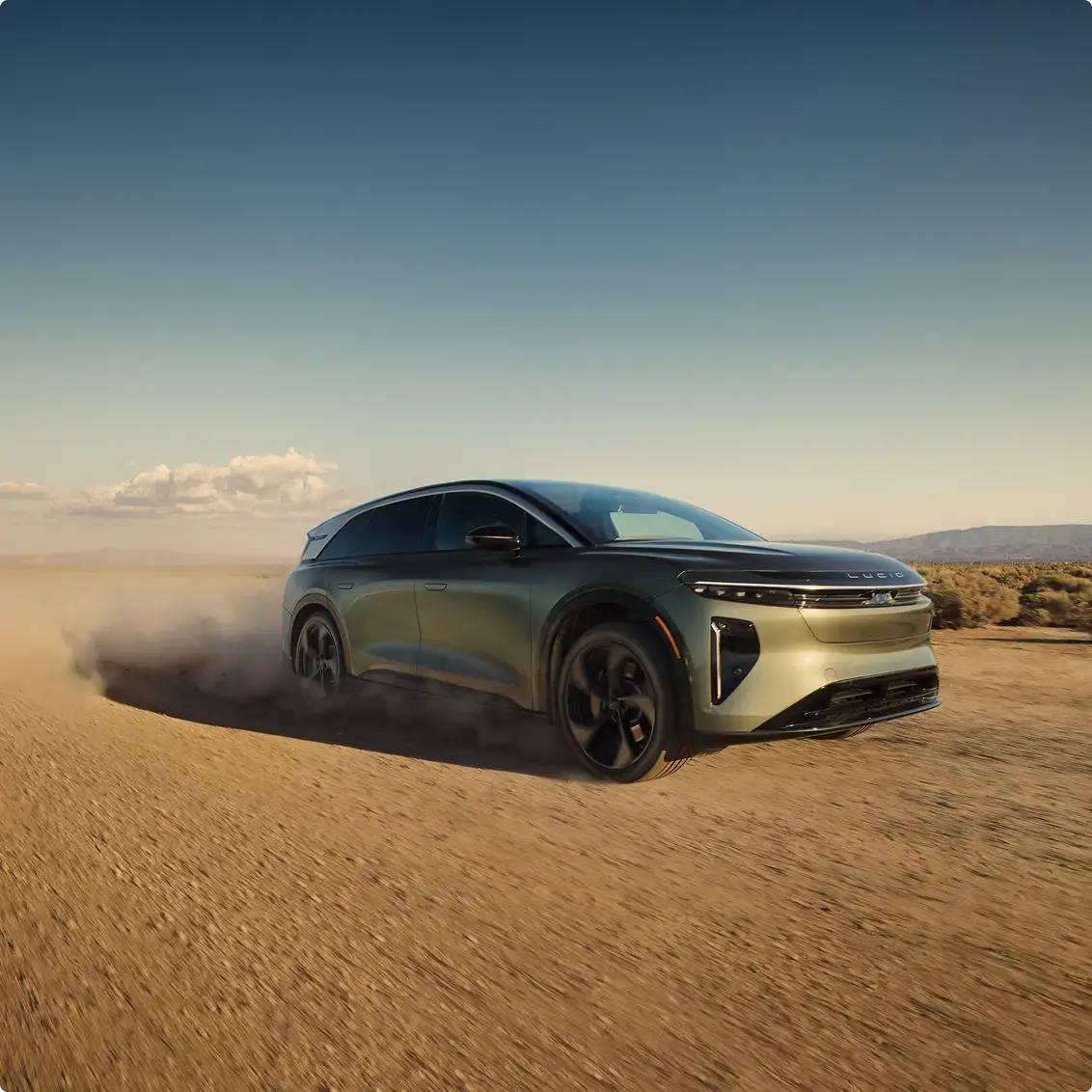 A Lucid electric SUV drives fast across a dusty desert terrain, kicking up a cloud of dust behind it.
