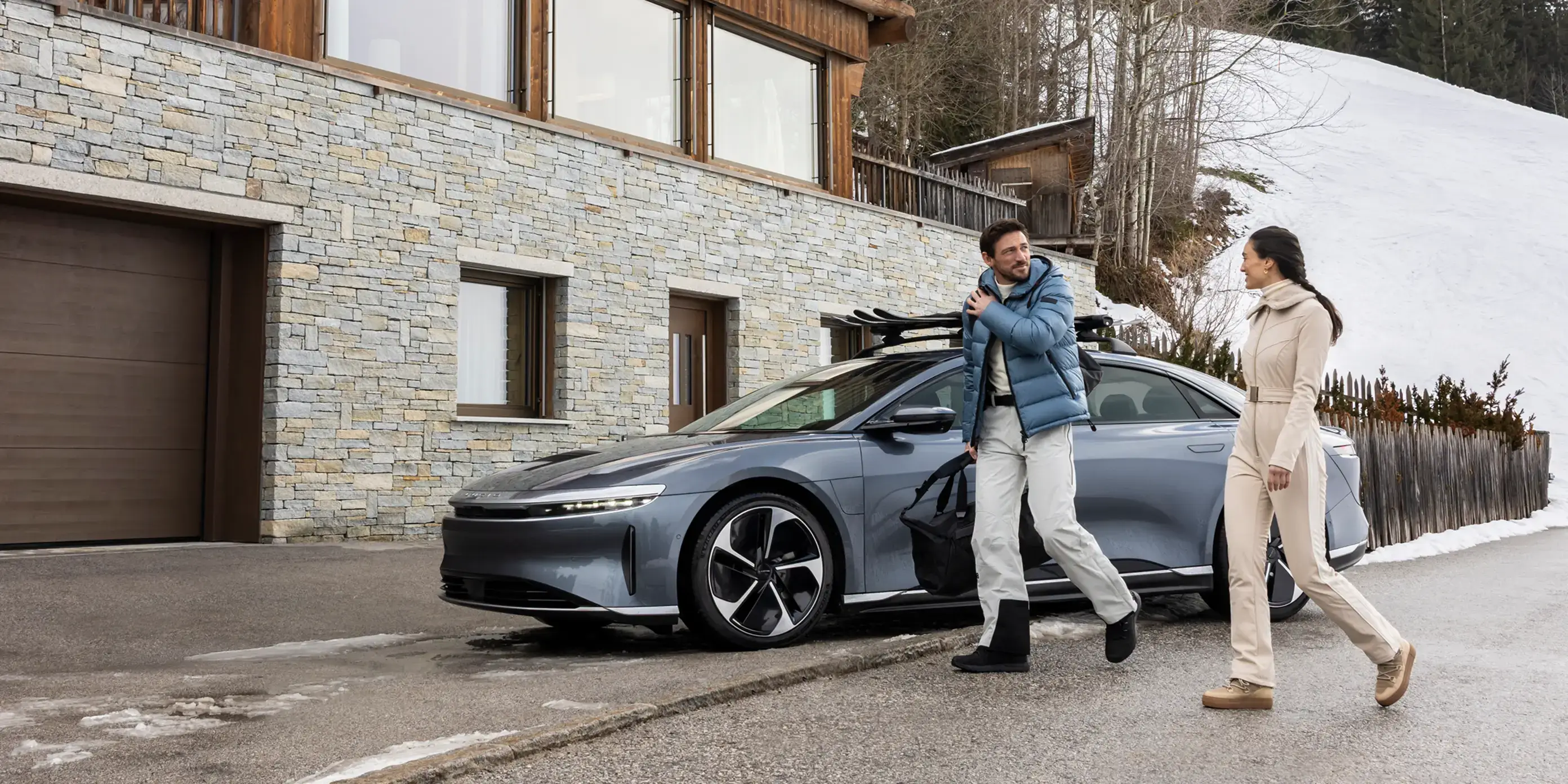 Deux personnes marchant près d'une Lucid Air équipée d’un porte-skis et garée devant une maison de montagne moderne en pierre et en bois.
