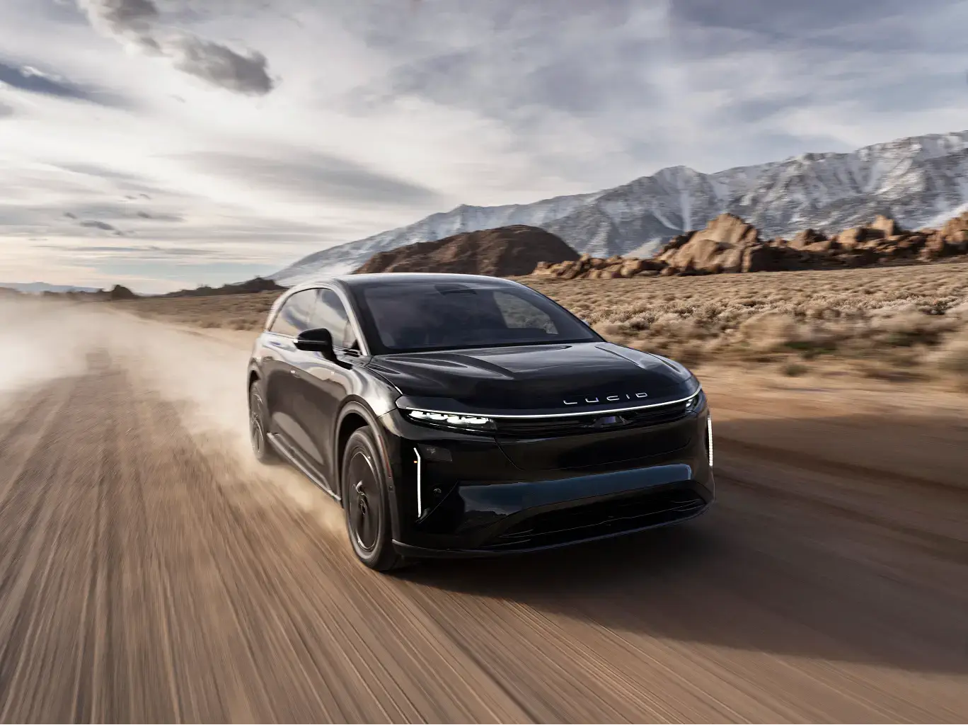 Black Lucid electric SUV speeding along a dusty desert road with snow-capped mountains in the background.