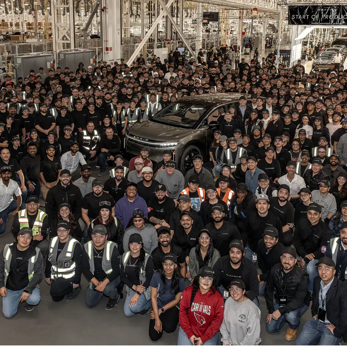 Large crowd of factory workers gathered around a new Lucid electric vehicle in a modern manufacturing facility.