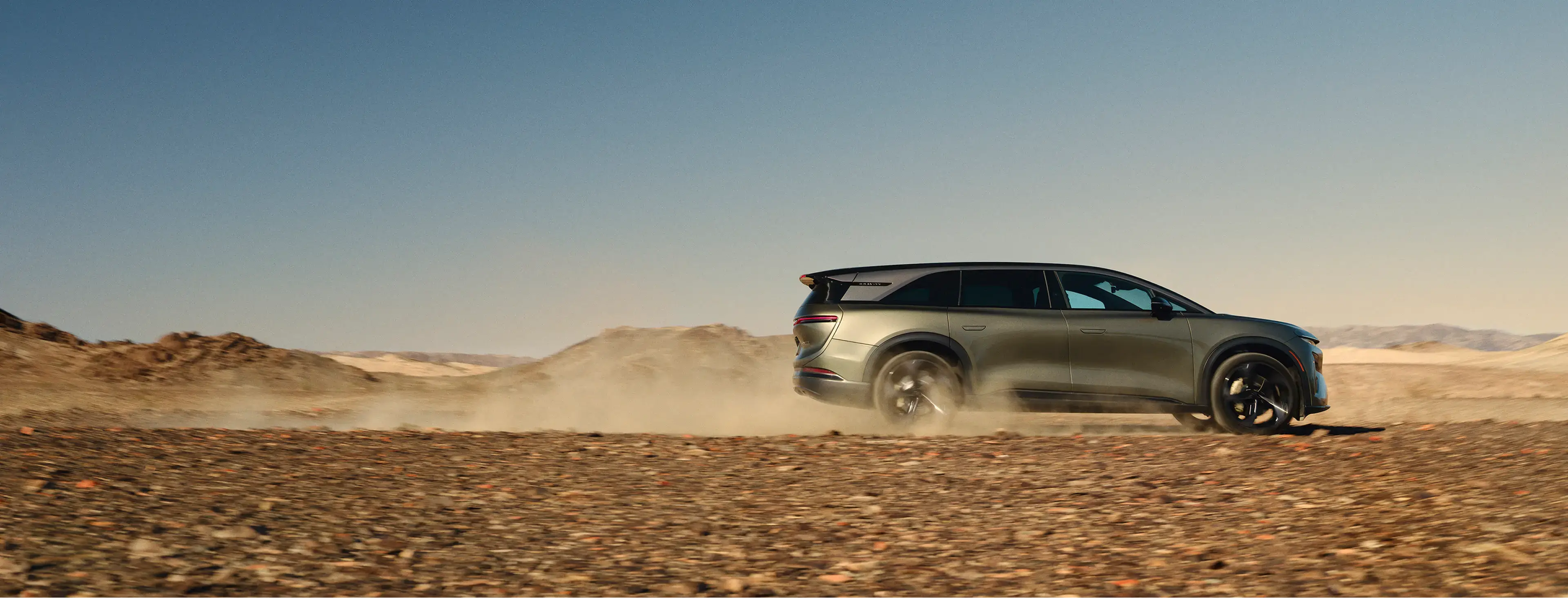 Lucid electric SUV driving across snowy terrain, kicking up powder with forest backdrop under blue sky.
