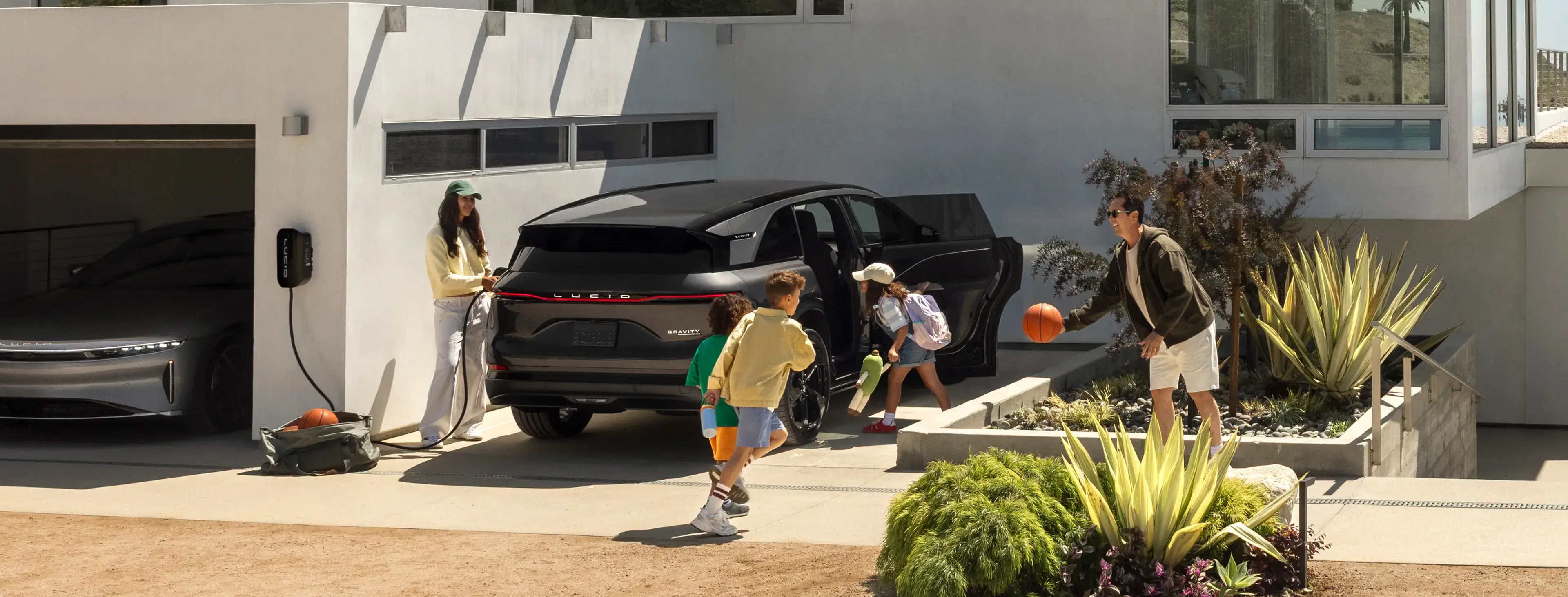 A woman charges a black Lucid Gravity SUV at home while two children load into the car in a sunny driveway.