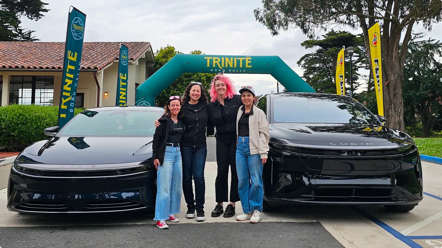 Four women posing between two black Lucid cars under a teal Trinité Road Rally inflatable arch outdoors.