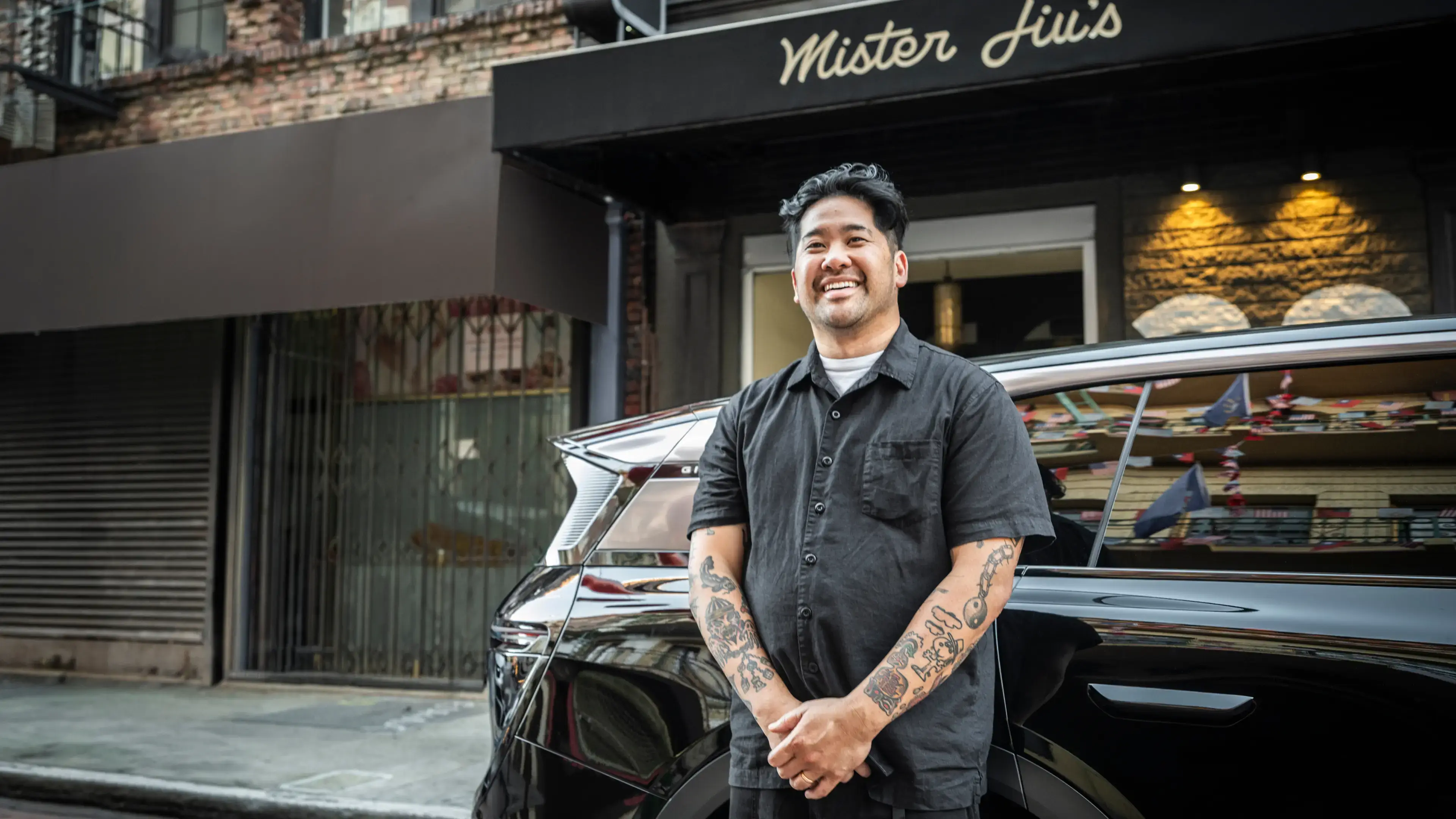Smiling tattooed man in a black shirt standing outside Mister Jiu's restaurant with hands clasped.