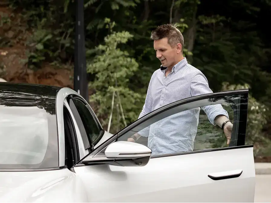 A smiling man in a blue striped shirt opens the door of a white car outdoors with green trees in the background.