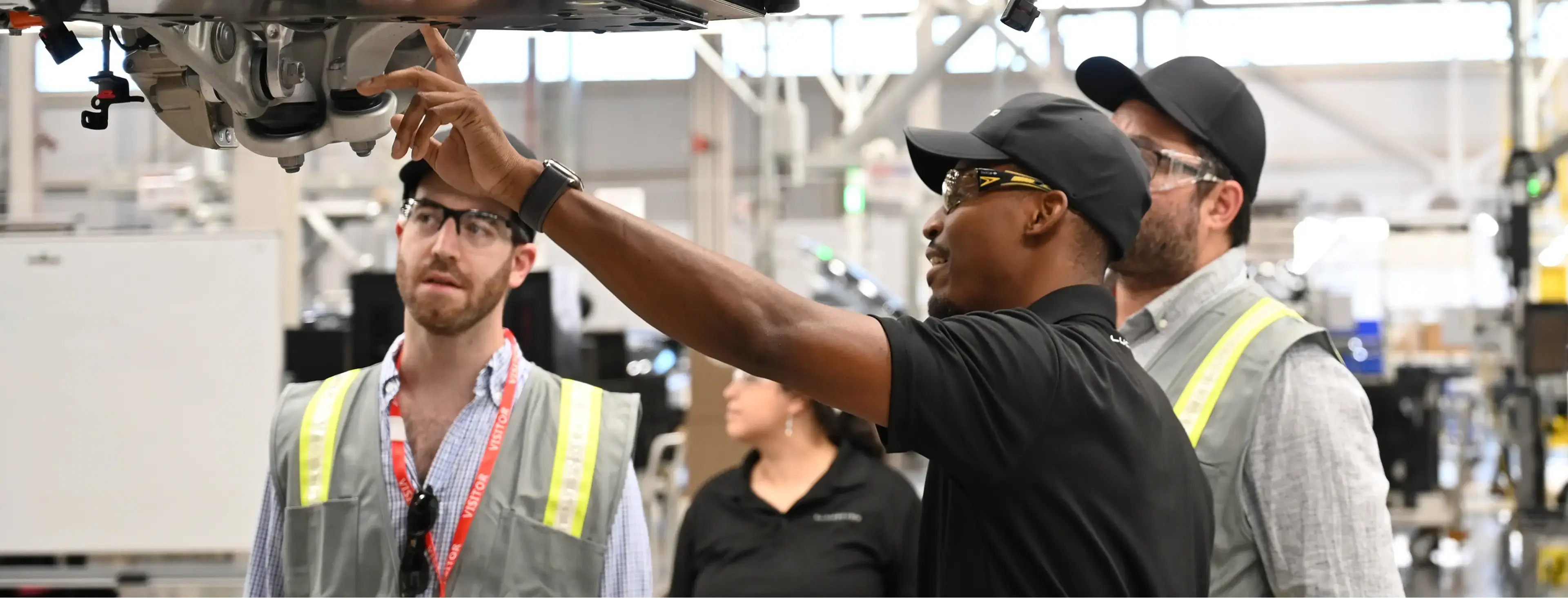 Workers in safety vests examine the underside of a vehicle on an assembly line in a manufacturing facility.