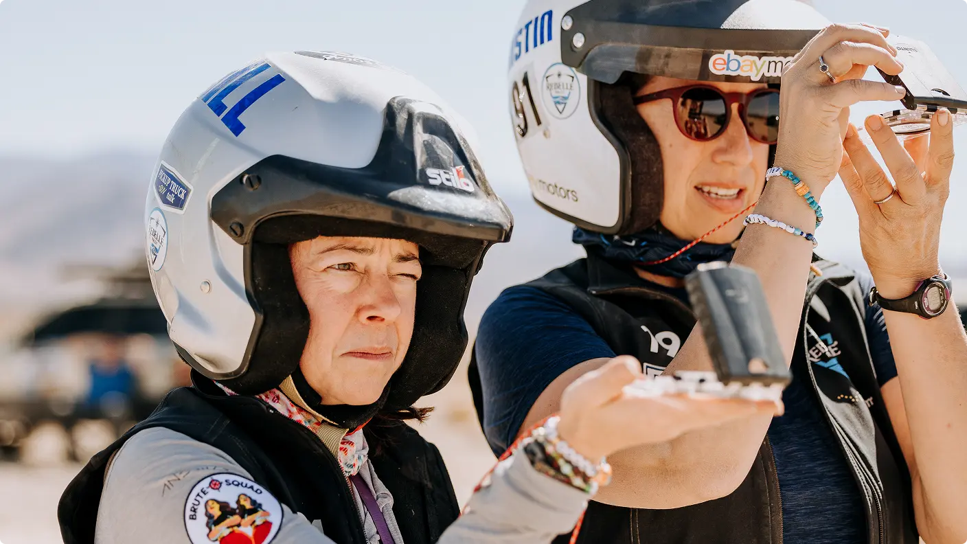 Two women wearing racing helmets examine a small device outdoors, with a desert landscape in the background.