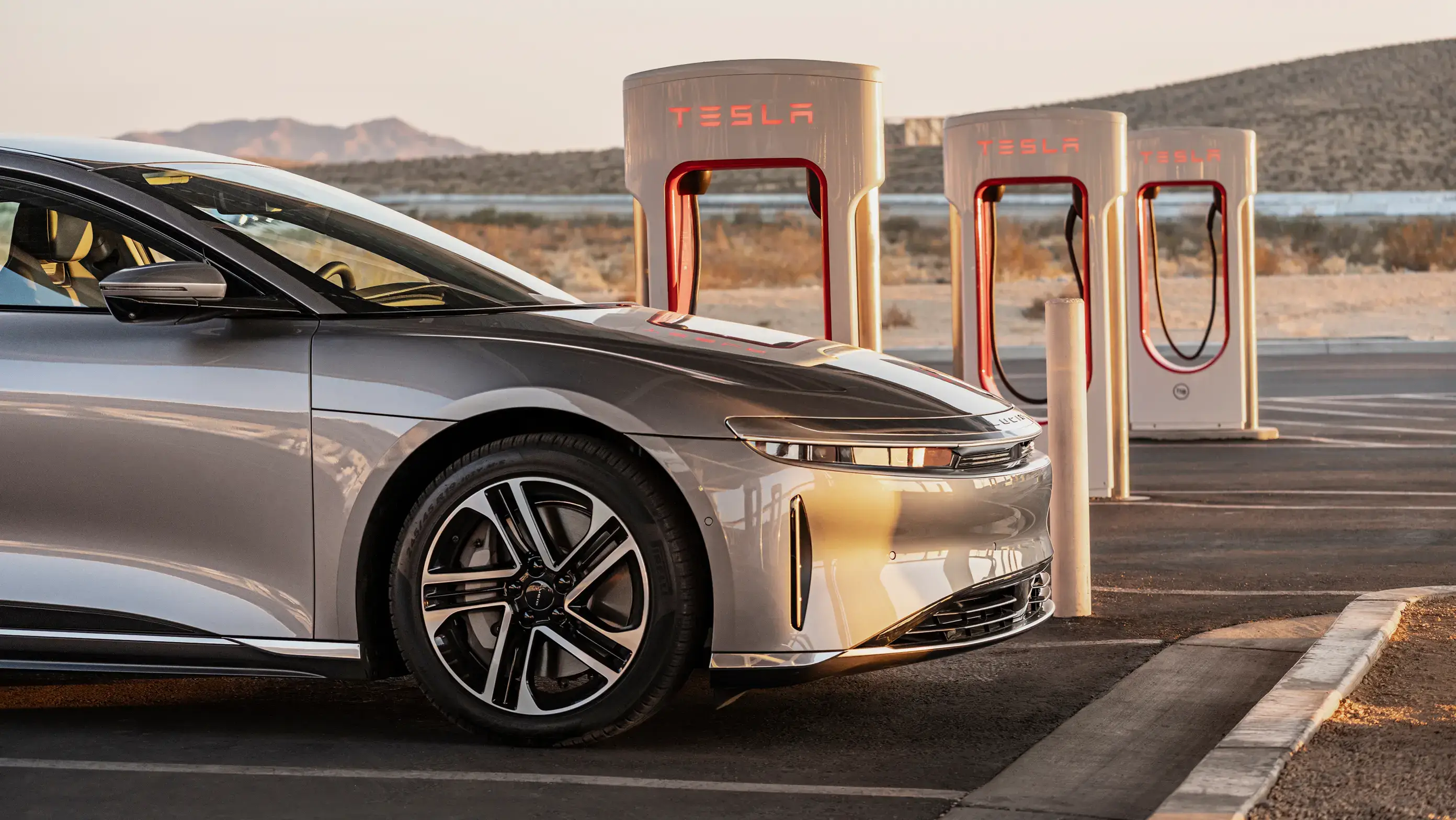 Lucid Air parked at Tesla Supercharger station in desert landscape with mountains in background.