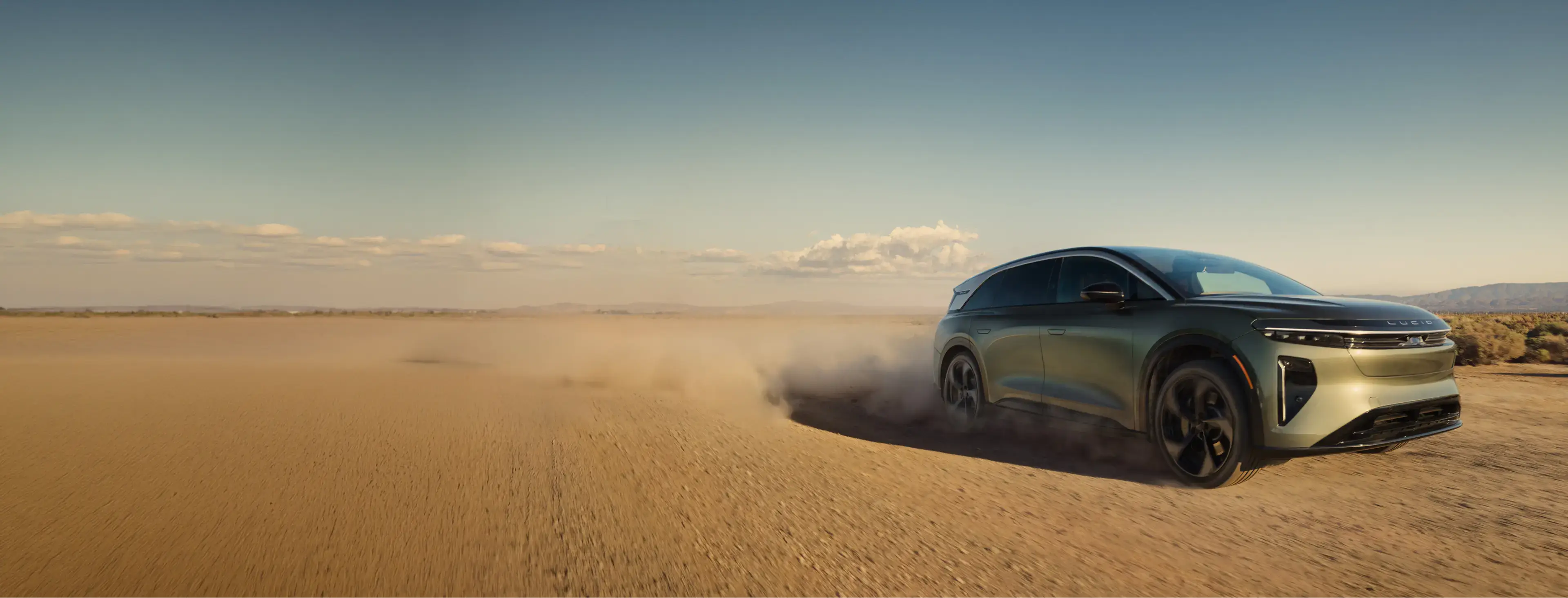 Lucid Gravity SUV driving across desert terrain, kicking up dust clouds against a clear blue sky and distant mountains.