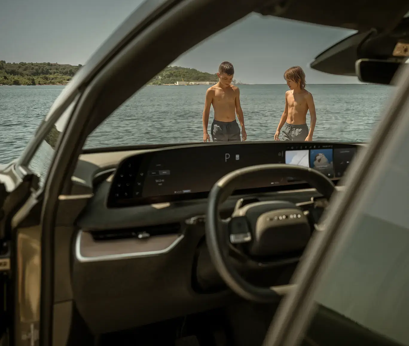 View from inside a car's interior showing two shirtless boys standing near a calm blue lake on a sunny day.