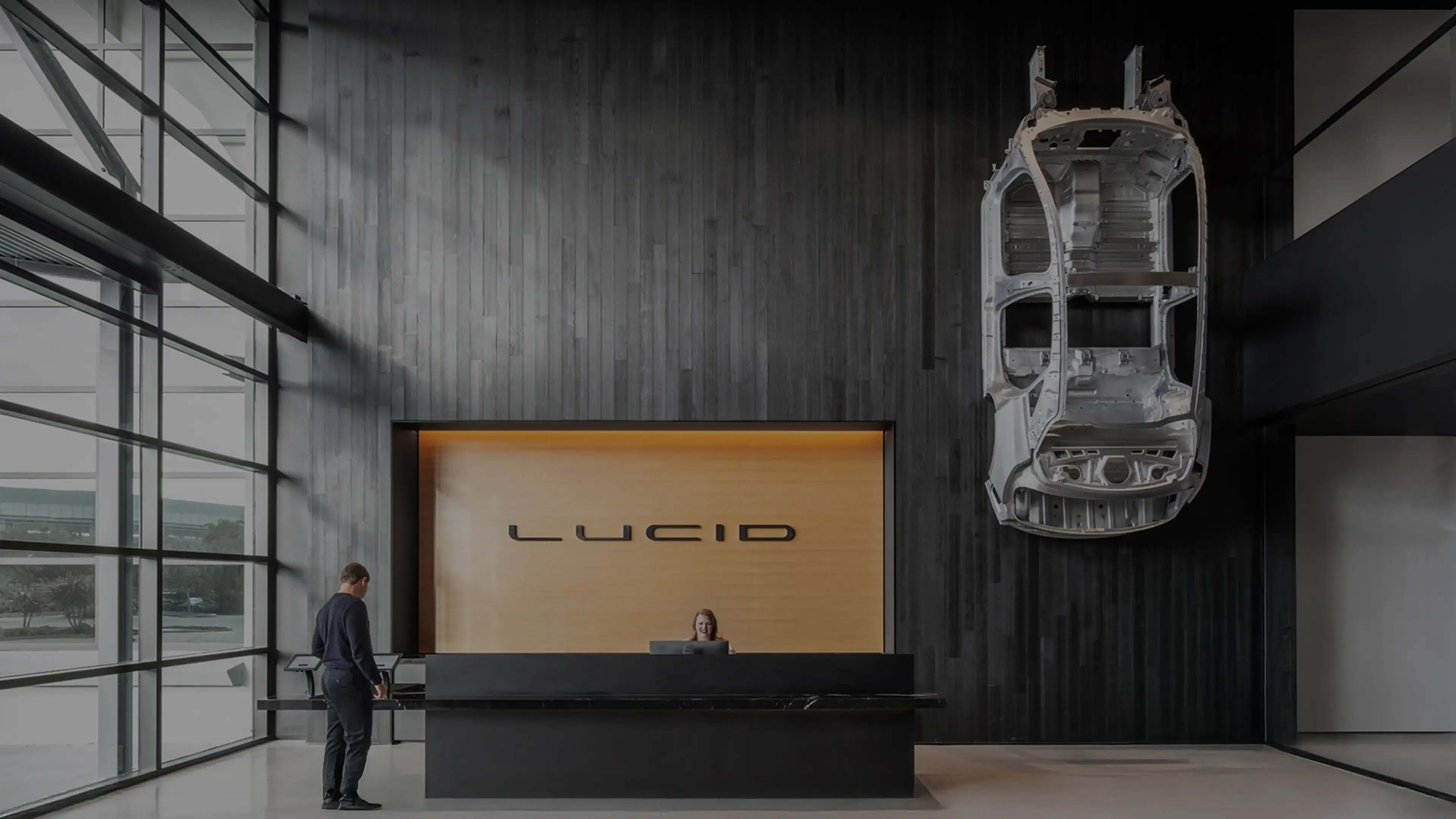 Modern Lucid Motors reception area with black desk, wooden wall display, and car chassis mounted as artistic installation.