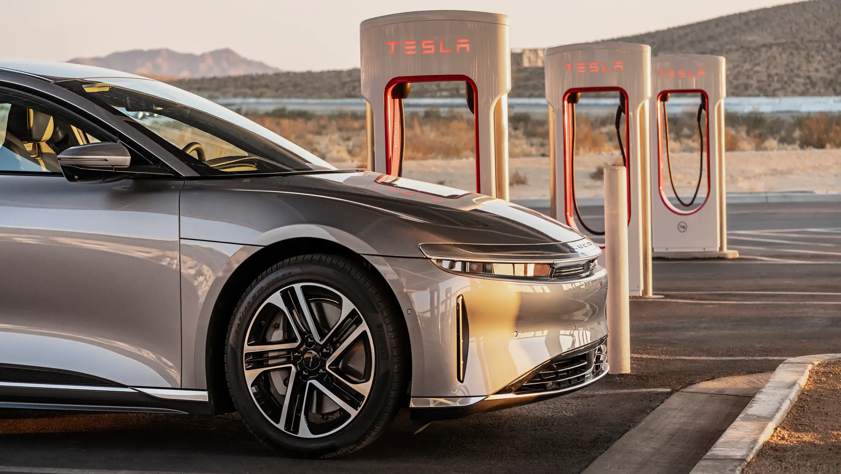 Lucid Air vehicle parked at Tesla Supercharger station with multiple charging units in desert landscape at dusk.