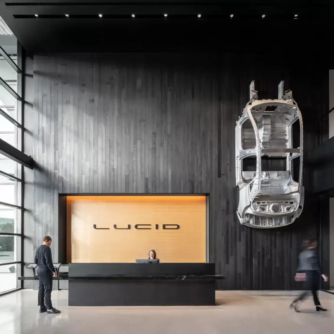 Modern Lucid Motors showroom with sleek black reception desk, wooden accent wall, and car chassis displayed as wall art.