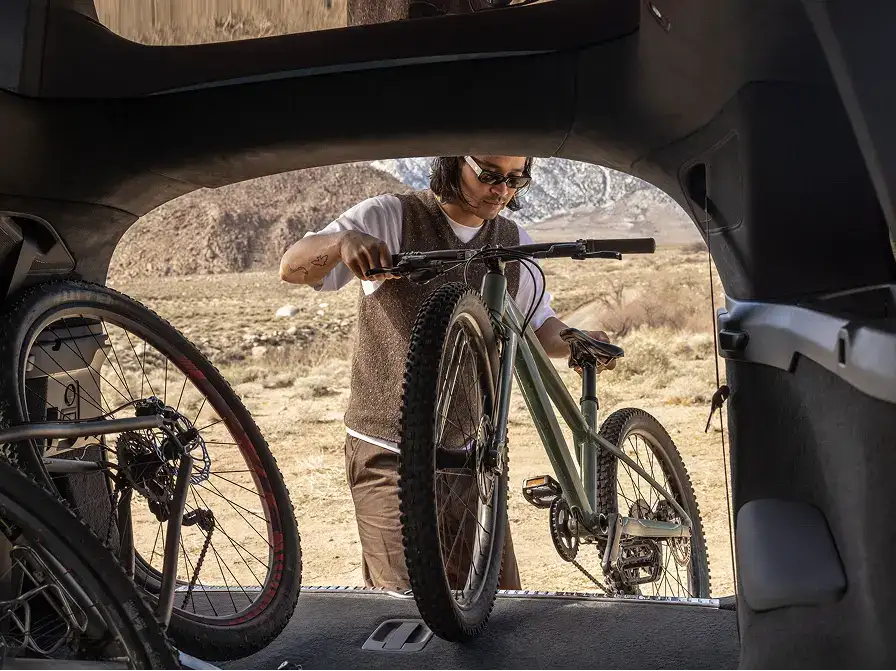 A person loading a green mountain bike into a vehicle's trunk, with a desert landscape visible in the background.