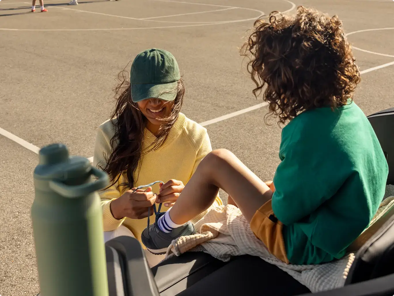 A woman and child sitting in a frunk; one in a yellow sweater and green cap ties the other's shoe, both smiling outdoors.