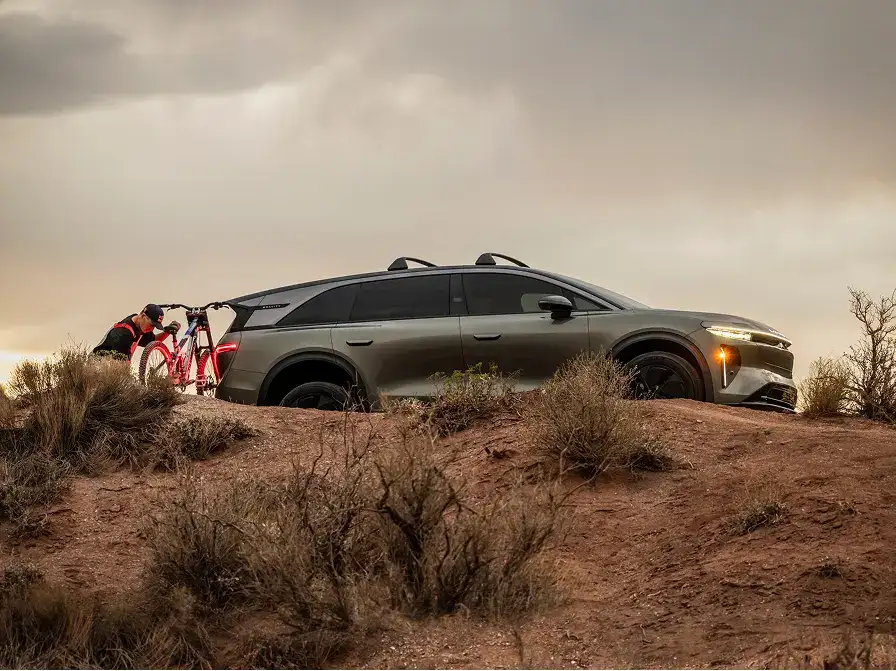 A Lucid SUV parked on red rocky terrain at dusk, with a person unloading a red bicycle from a rear bike rack.