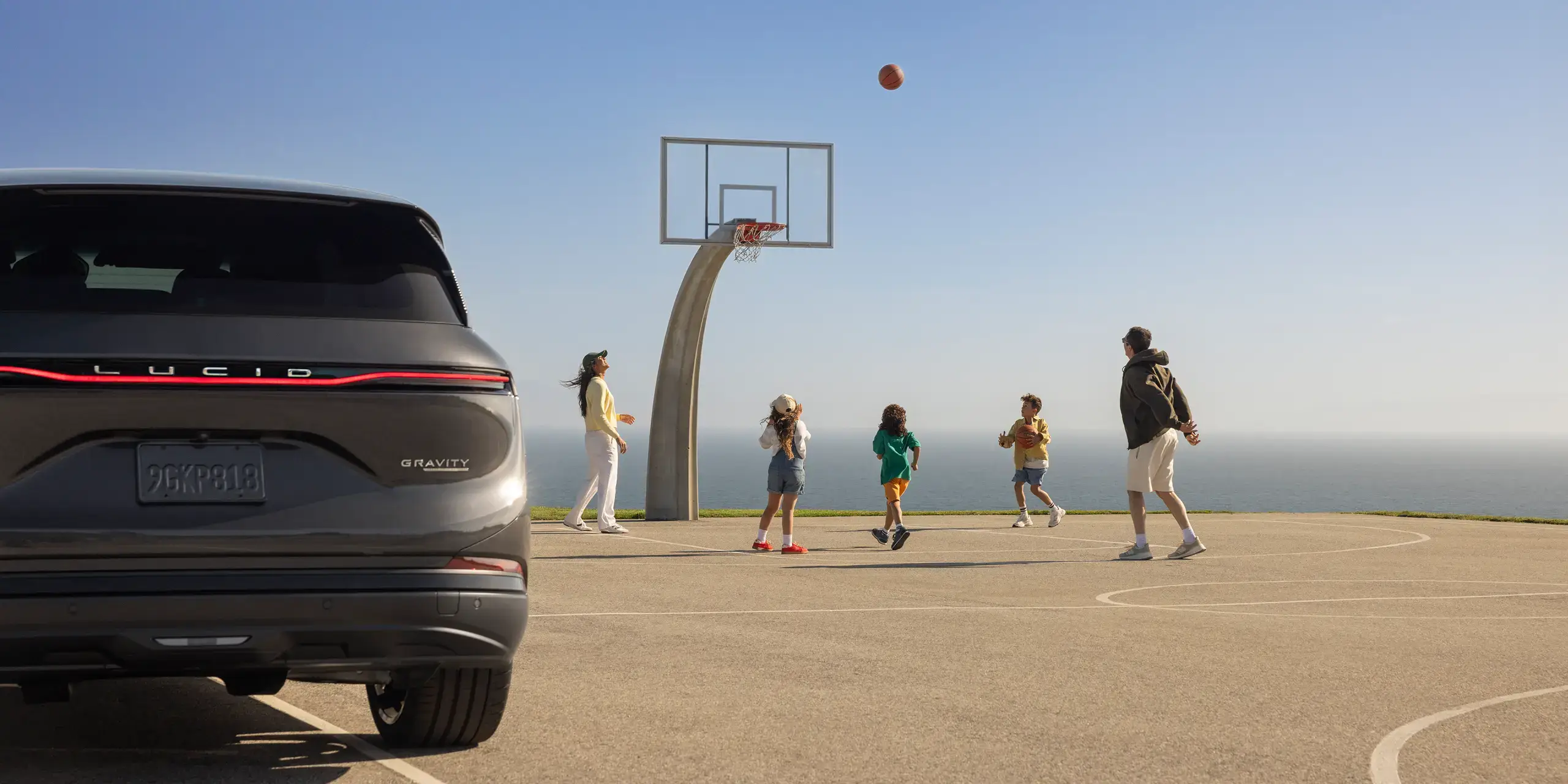 A family plays basketball on an outdoor court near the ocean, with a gray Lucid Gravity SUV parked in the foreground.