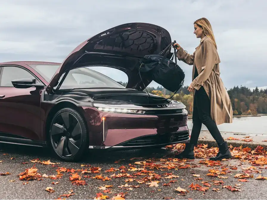 Woman in a beige coat loading a bag into the frunk of a dark red Lucid Air sedan surrounded by autumn leaves.