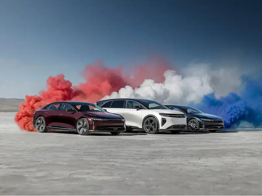 Three Lucid electric cars in dark red, white, and dark blue, posed outdoors with matching red, white, and blue smoke clouds behind them.