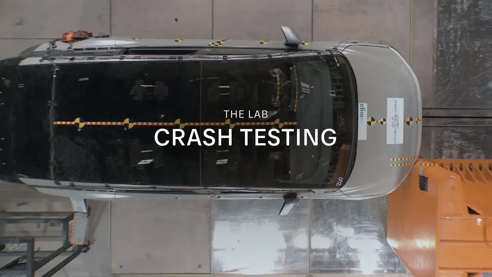 Overhead view of a vehicle in a crash test facility with "THE LAB CRASH TESTING" text overlaid on the image.