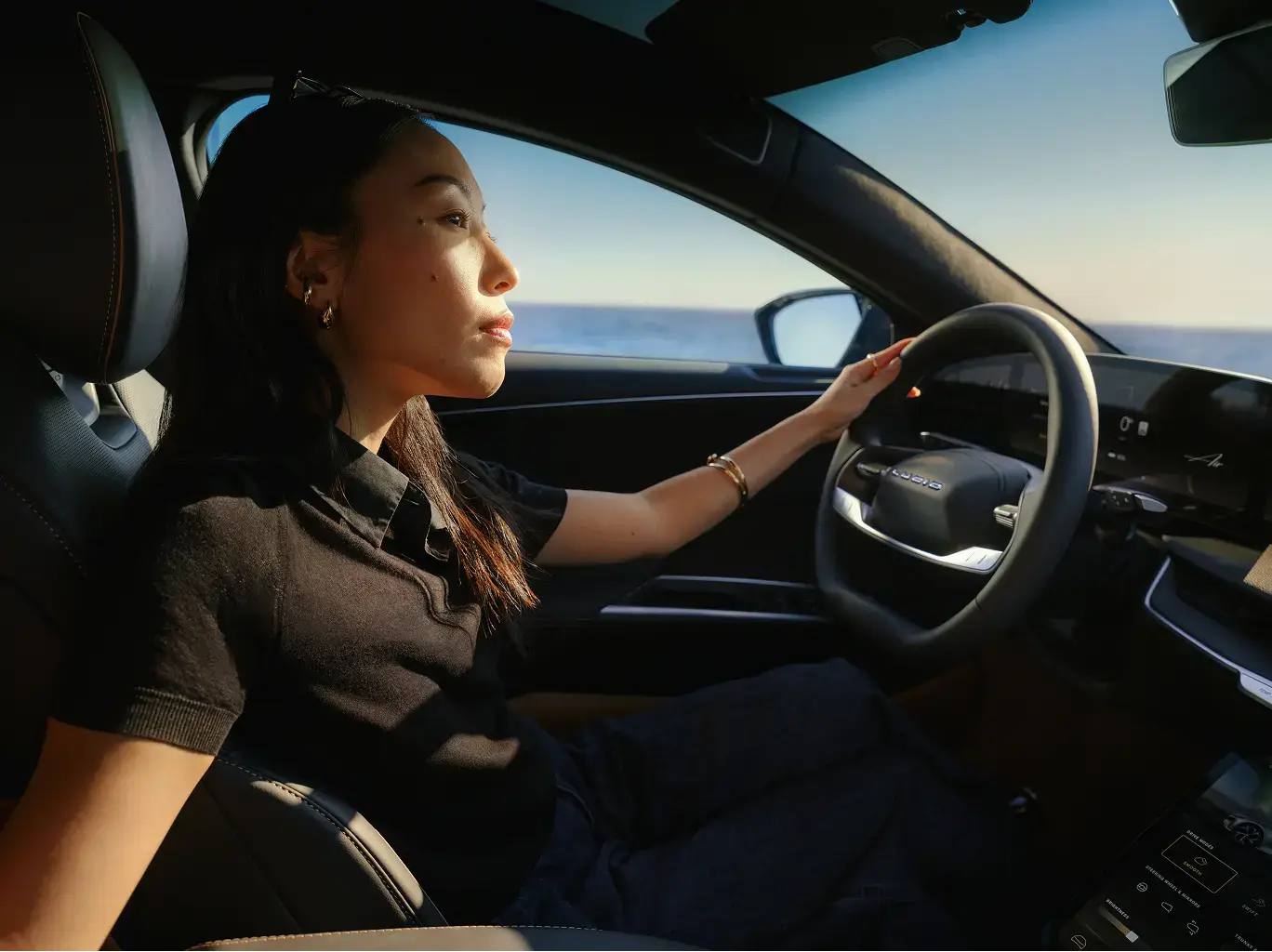 Person driving a car along coastal road, illuminated by golden sunlight, with ocean visible through the window.