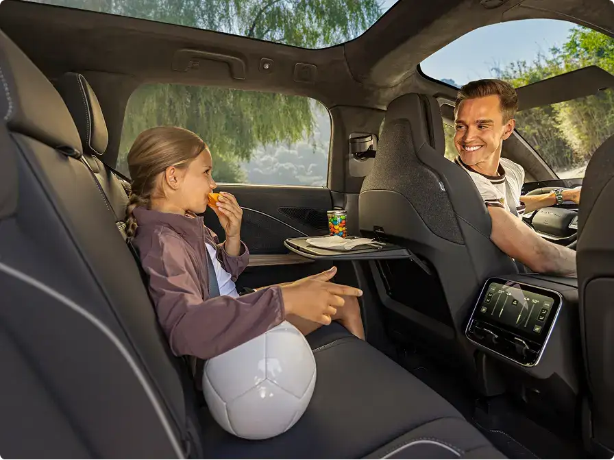 A smiling man driving an SUV turns to a girl in the backseat eating a snack, with a soccer ball and rear touchscreen visible.