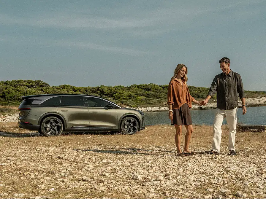 A couple holds hands near a sleek gray SUV parked on a rocky coastal landscape with water and greenery behind them.