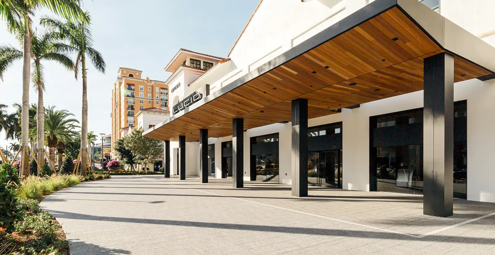 Lucid Motors showroom exterior with a wood-paneled canopy, black columns, glass storefront, palm trees, and a sunny Florida plaza.