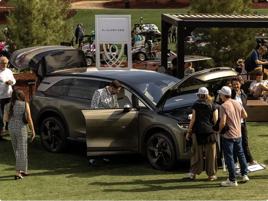 People examining a dark green Lucid SUV at an outdoor auto showcase event, with a Lucid branded sign displayed nearby.