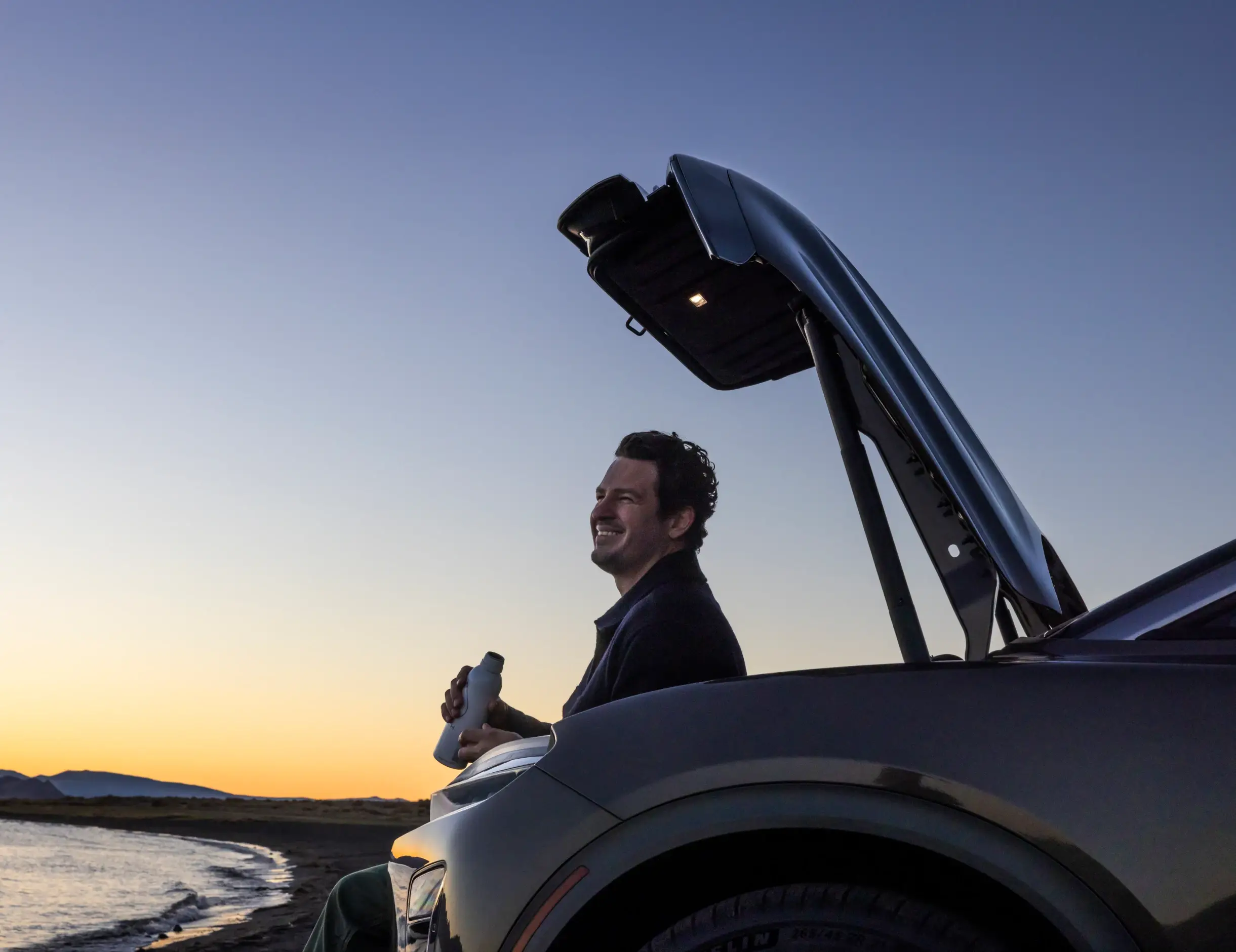 Een persoon zit bij de open achterbak van een Lucid Gravity tijdens zonsondergang, met een drankje in de hand terwijl hij uitkijkt over het water.
