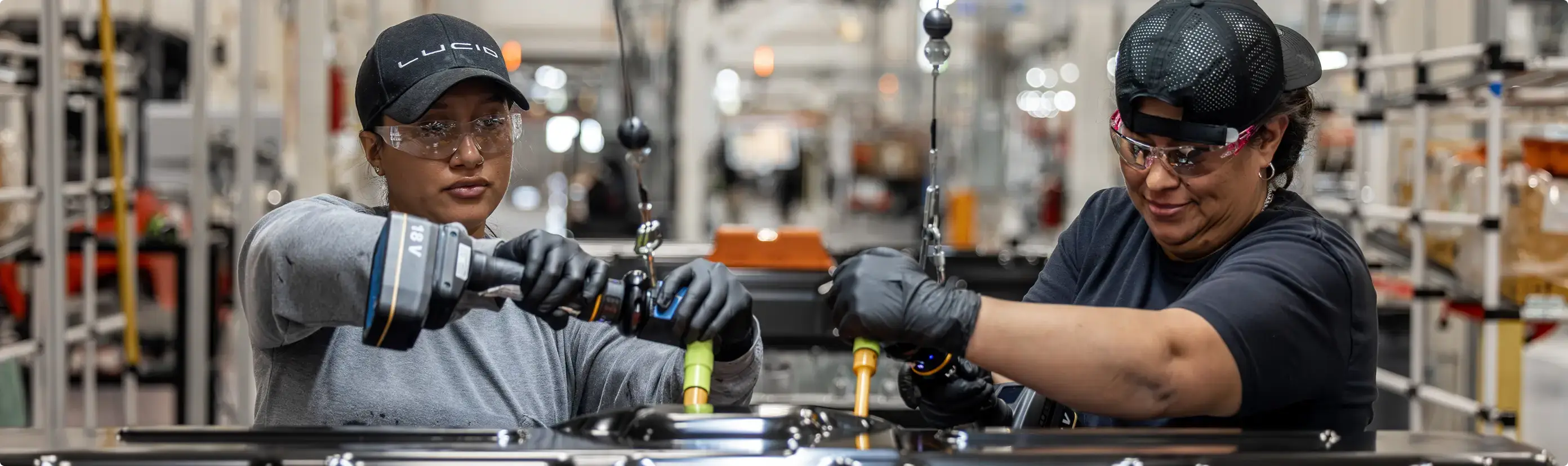 Two factory workers wearing safety glasses and gloves assembling components on a manufacturing line.