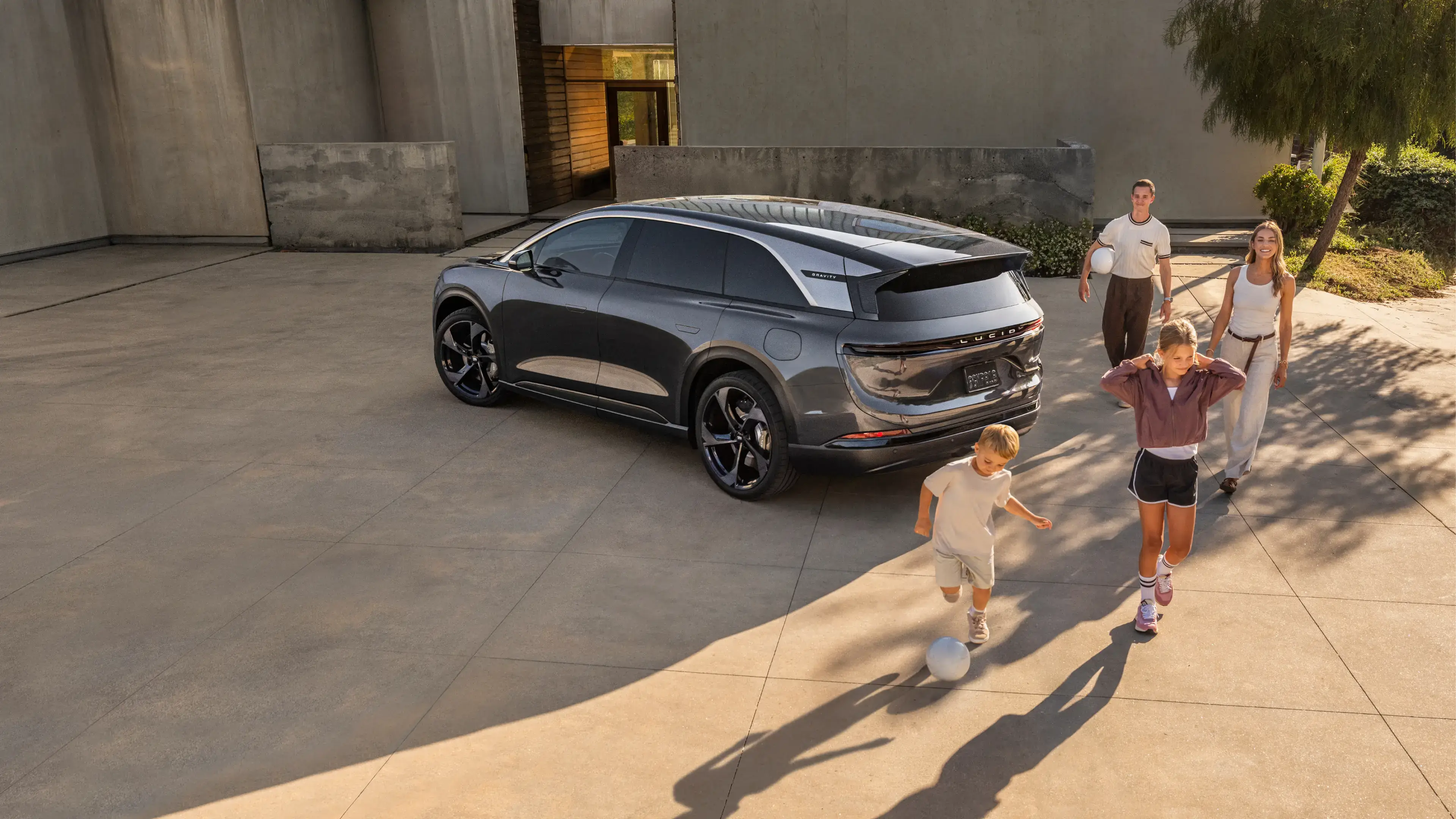 A family plays soccer near a dark gray Lucid Gravity SUV parked on a sunlit concrete driveway.