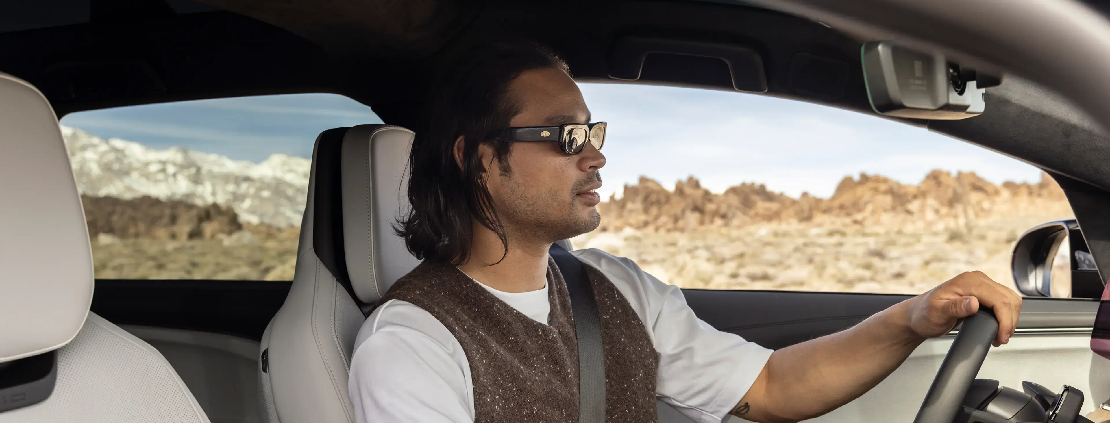 Man with sunglasses and long hair driving a car through a desert landscape with rocky terrain and mountains.