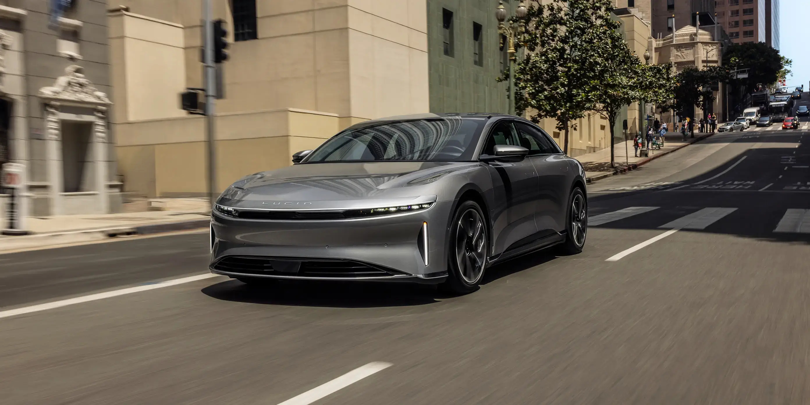 A silver Lucid Air electric sedan driving through a sunny urban street with buildings and pedestrians in the background.