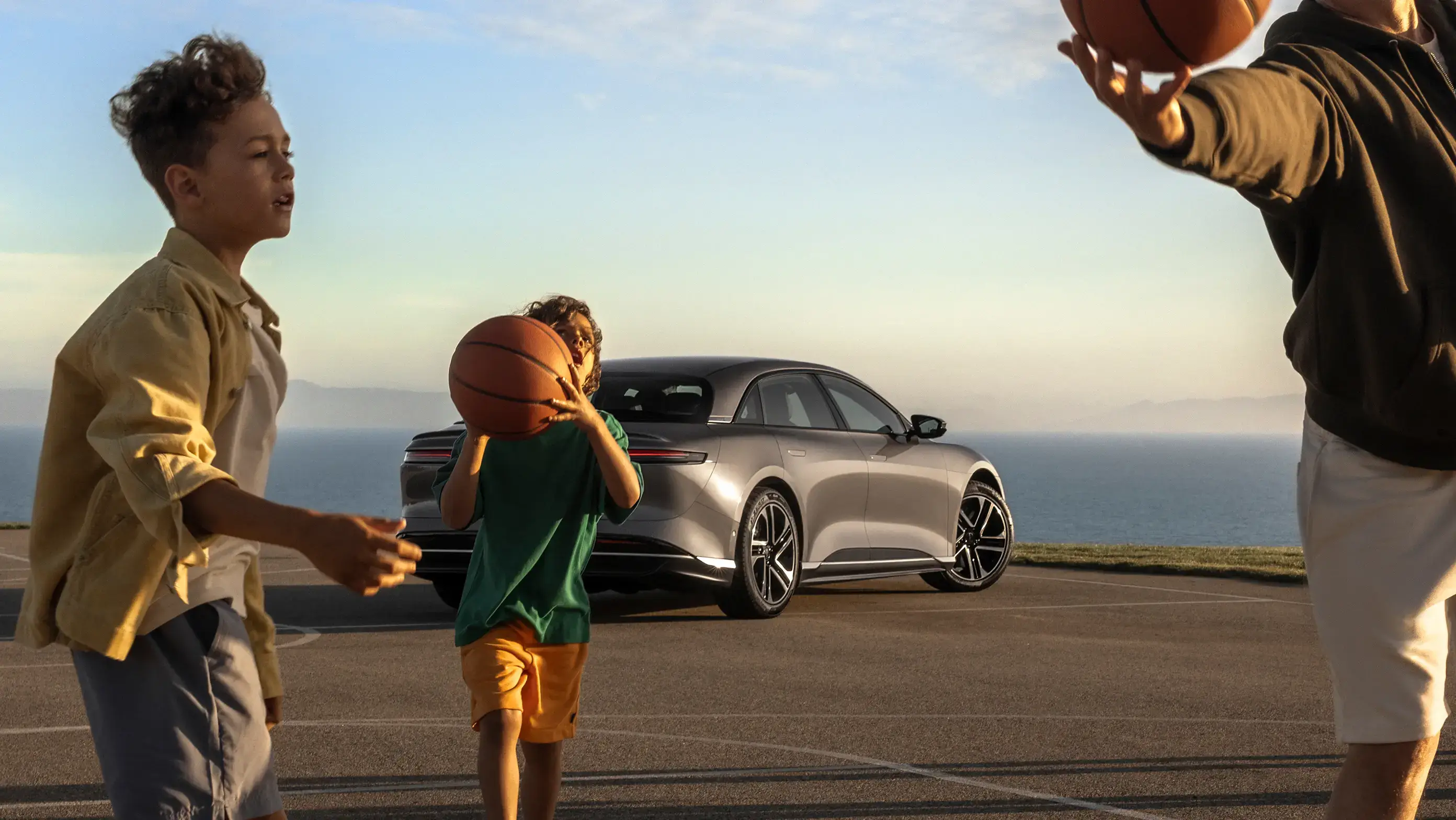 Two children playing basketball outdoors near a Lucid Air sedan parked by the ocean at sunset.