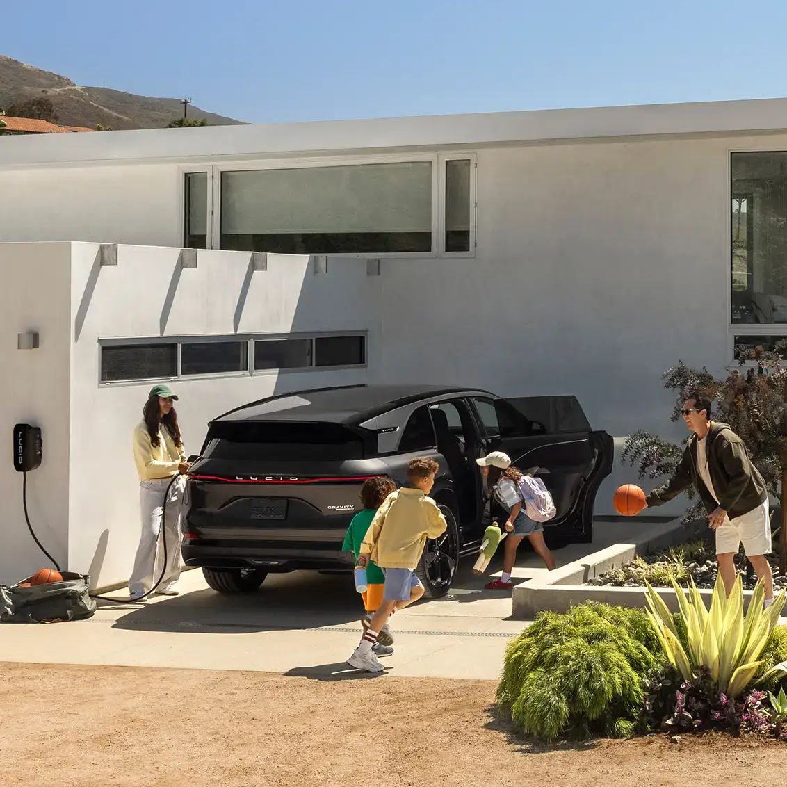 A family outside a modern home with a black Lucid Gravity SUV charging, two kids playing, and a man holding a basketball.