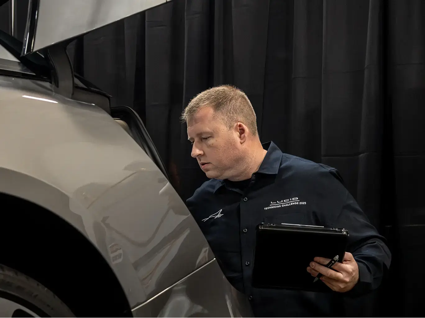 Technician in Lucid uniform inspecting a silver vehicle while holding a tablet and pen against a black backdrop.