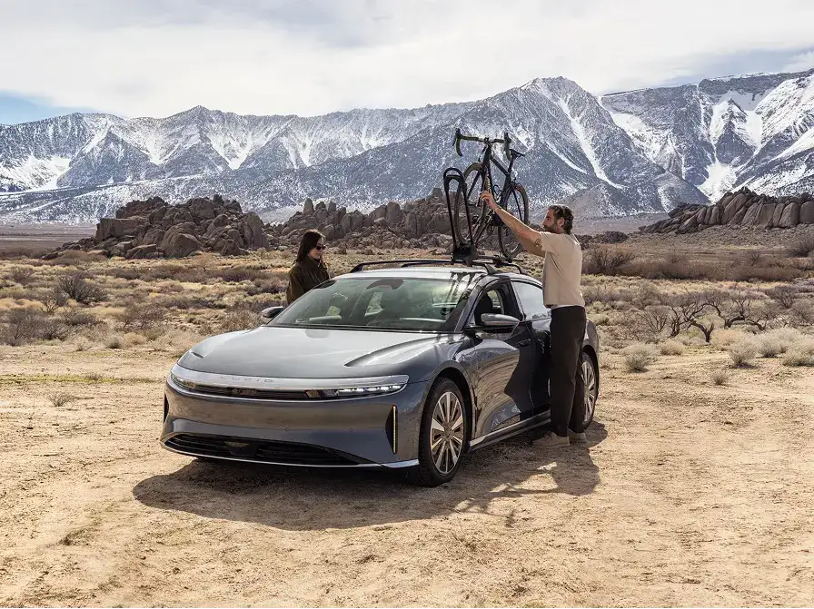 Two people load a bicycle onto a Lucid Air's roof rack in a desert landscape with snow-capped mountains behind them.