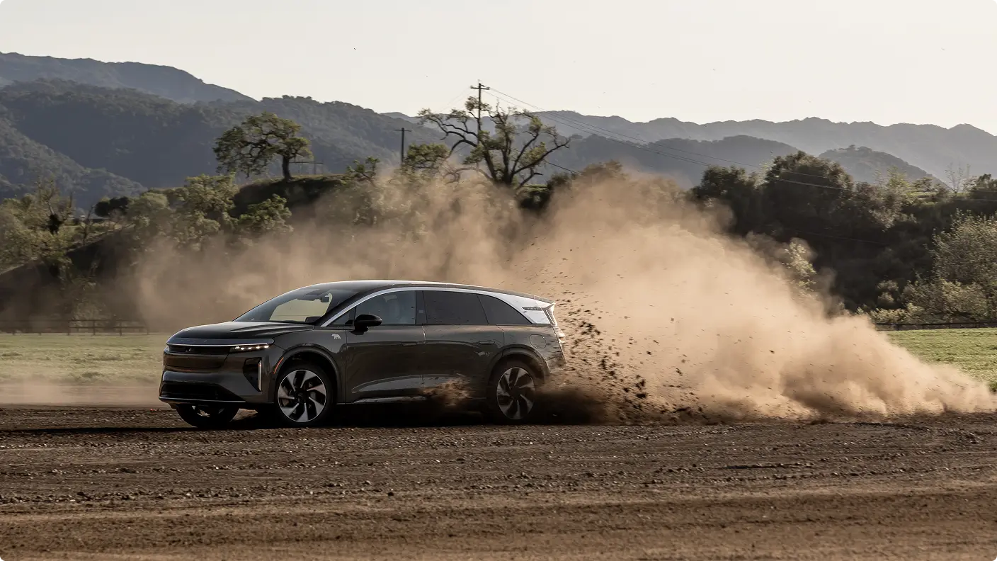 A Lucid Gravity SUV drives aggressively on a dirt surface, kicking up a large dust cloud, with mountains and trees in the background.
