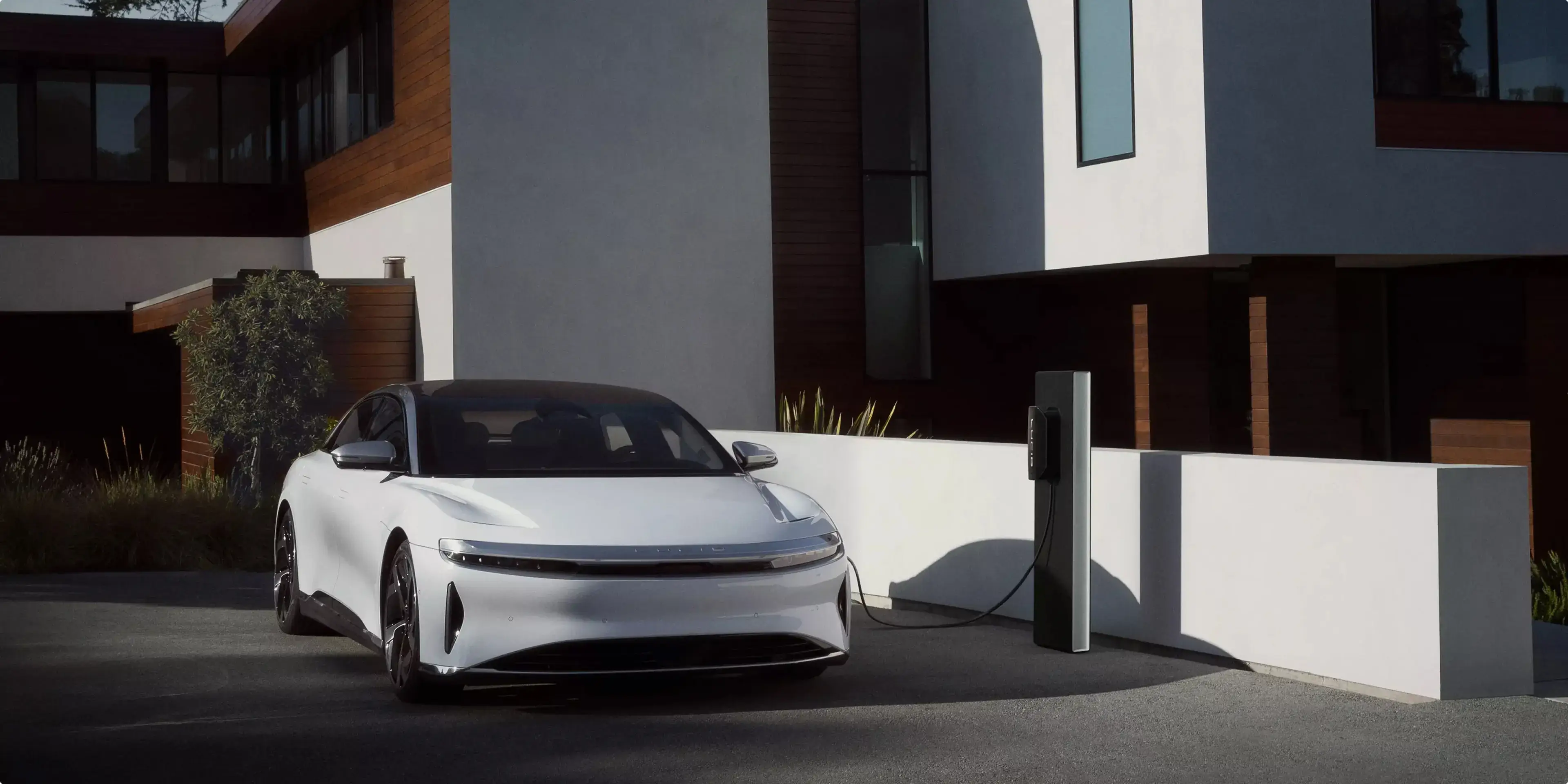 Voiture électrique Lucid Air en recharge devant une maison moderne avec panneaux extérieurs gris et en bois et une architecture minimaliste.